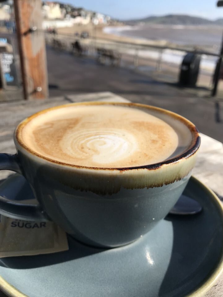 Craving normality. Image shows a cup of coffee, a latte, with a beach blurred out on the background