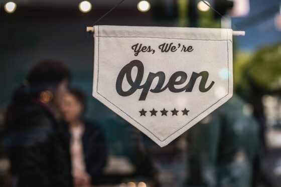 Image shows a sign saying "Yes, we're open" with a blurred image of two people in the backgroun