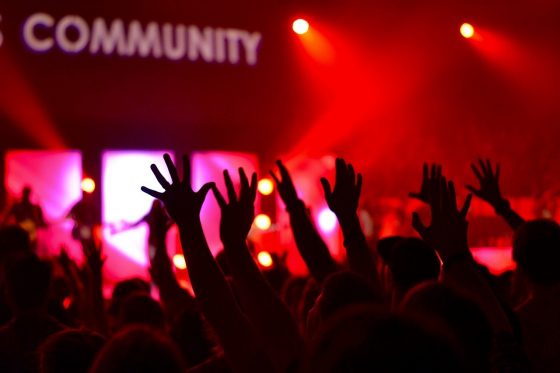 Image shows people with their hands in the air inside possibly a night club with the word "community" picked out in lights.