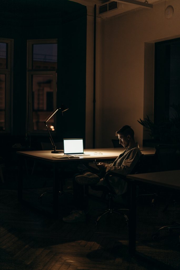 Image shows a bright laptop screen in a relatively dark room. We can just make out a man sitting in a chair.