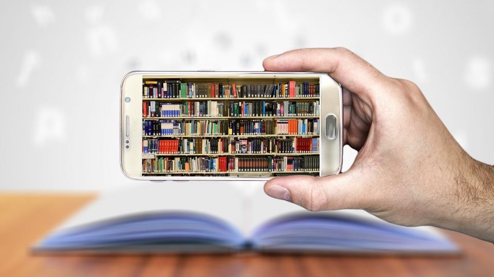 Image shows an image of a library of books on a smartphone screen with a real book on a table in the background
