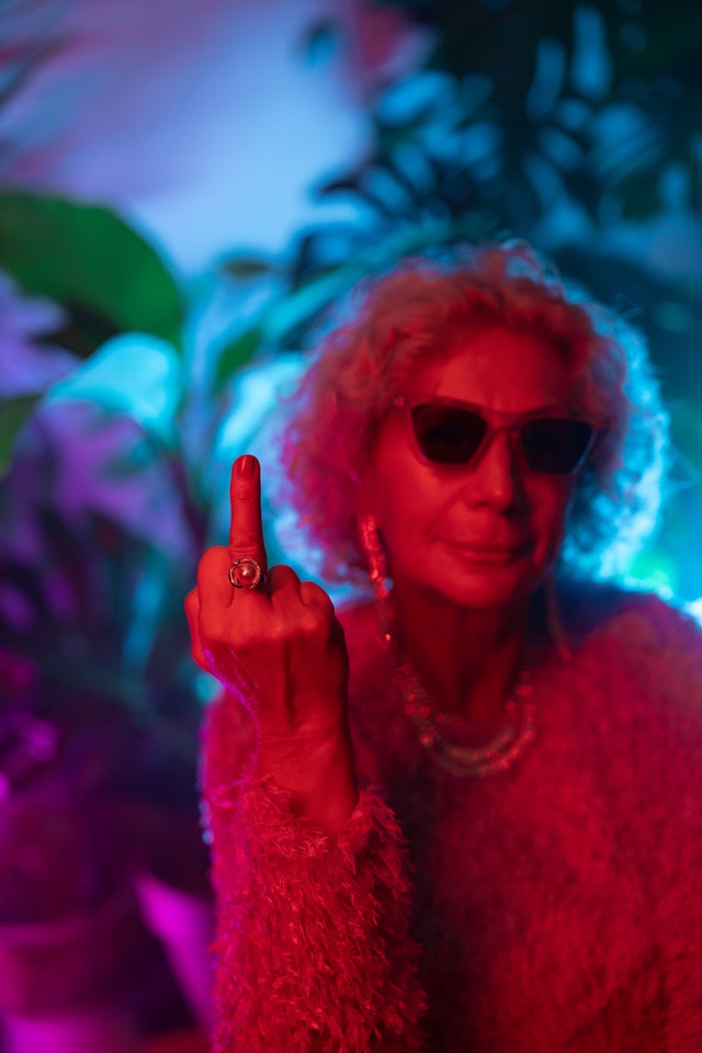 Image shows an older woman with sunglasses on flipping the middle finger