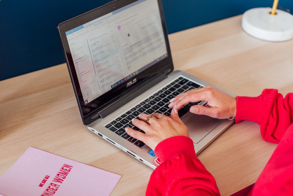 Image shows women's hands typing on a laptop keyboard. Red jumper. Pink notebook. White lampbase | Helen Tarver Freelance Copywriter