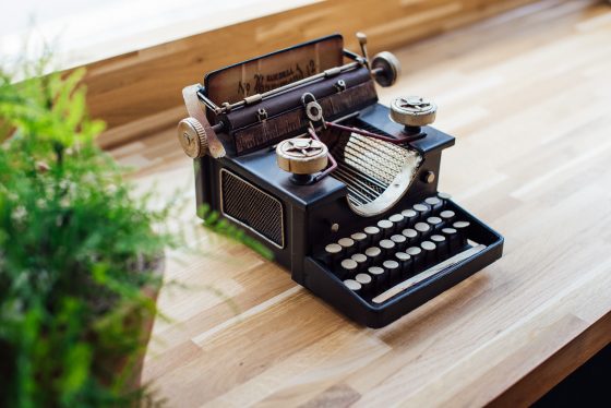 Image shows a vintage looking stenography machine or small typewriter on a wooden background with a green plant to the left side. A technology that is dated and in need of a refresh.