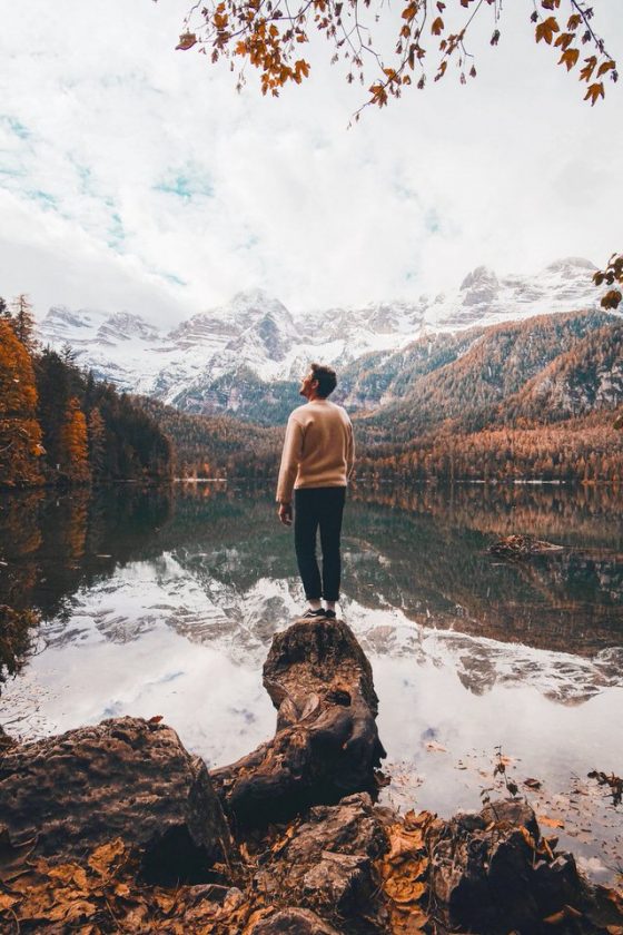 Image shows a person looking up at snow covered mountains. The lake seems perfectly still so the reflection is almost mirror like.