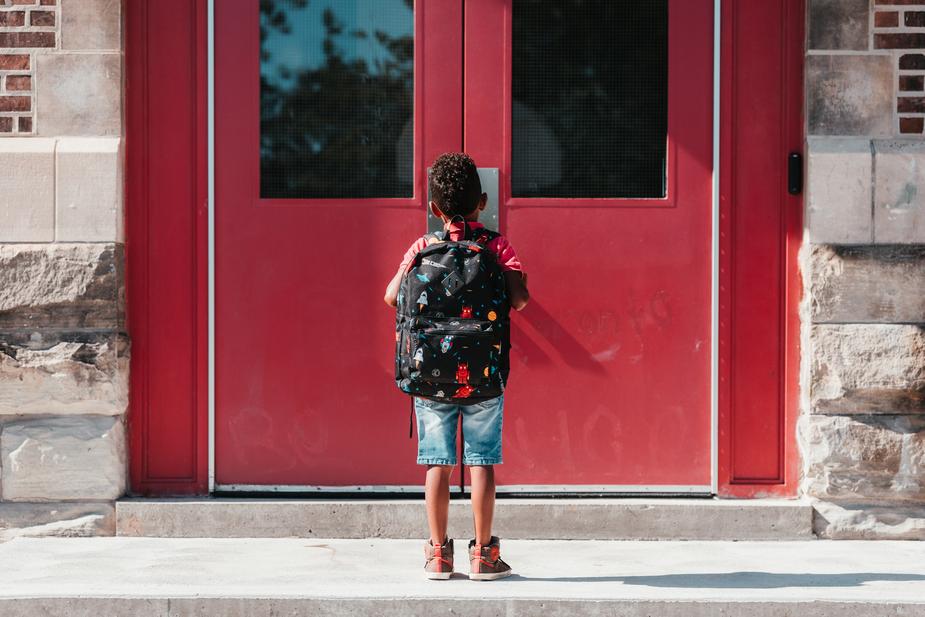 Image shows a small child in blue shorts outside a red school door. Learning is lifelong.